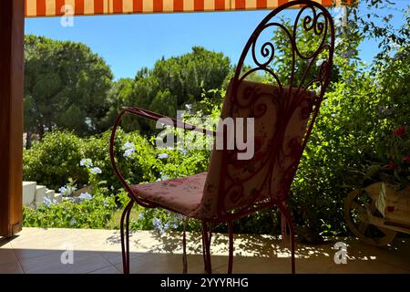Klassischer, schmiedeeiserner roter Stuhl und rosafarbene Kissen im alten Stil auf der Terrasse der Ferienvilla im Garten, Bäume, Blumen, blauer Himmel Stockfoto