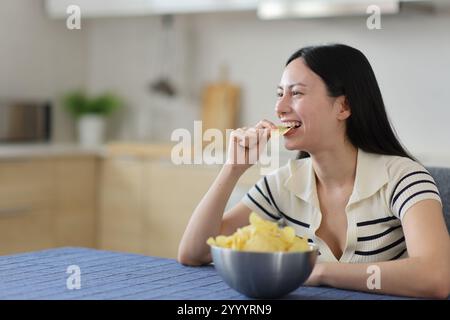 Glückliche asiatische Frau, die Kartoffelchips isst und zu Hause in der Küche wegschaut Stockfoto