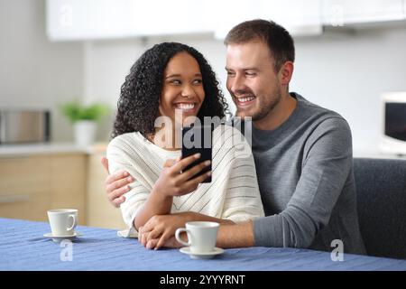 Glückliches interrassisches Paar, das telefoniert und sich zu Hause in der Küche ansieht Stockfoto