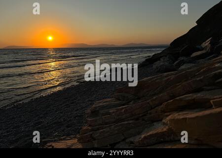 Sonnenuntergang an der Küste Ägäisches Mittelmeer, Sonnenschirme, Sonnenliegen, Sonnenuntergang hinter Bergen am Horizont, Reflexionen auf dem welligen Wasser Stockfoto