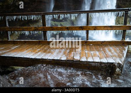 Ein Holzsteg, der mit Frost bedeckt ist, führt zu einem atemberaubenden Wasserfall. Eiszapfen hängen am Geländer und zeigen die eisige Schönheit des Winters in einem Naturpark. Stockfoto