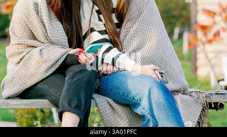 Zwei junge Frauen sitzen zusammen im Freien, in eine gemütliche Decke gehüllt, halten Smartphones und genießen einen zwanglosen Moment bei kühlem Wetter. Stockfoto