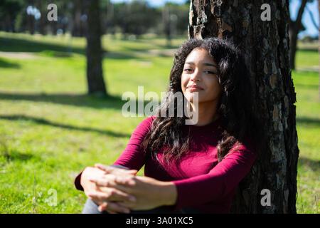 Eine ruhige junge lateinfrau genießt einen friedlichen Moment in der Natur, sitzt auf Gras und lehnt sich an einen Baum im Park Stockfoto