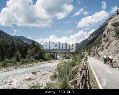 Ein gepflasterter Radweg entlang der Alpe Adria Radweg in Italien, umgeben von üppigen grünen Bergen und einem malerischen Fluss. Eine Brücke überspannt das Tal, während eine c Stockfoto