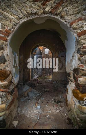 Durch ein bogenförmiges Portal, ein Blick in den Verfall: Eine vergessene Passage mit verwitterten Steinen und einer offenen Tür, die zur Erkundung einer vergangenen Ära einlädt. Stockfoto