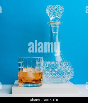 Eine wunderschön gefertigte Glasdekantierung hält Whiskey neben einem mit Eis gefüllten Kristallbecher, alles vor einem leuchtend blauen Hintergrund. Stockfoto
