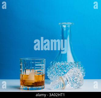 Knuspriger Whiskey, serviert auf Eis in einem Glas neben einer dekorativen Kristallkaraffe auf glatter Oberfläche. Die perfekte Wahl für anspruchsvolle Zusammenkünfte. Stockfoto
