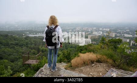 Frau Tourist steht auf dem Maschuk Berg mit Blick auf Pyatigorsk, Stavropol Krai, Russland. Der junge Mensch schaut von oben auf die nebelige Stadt. Reisekonzept p Stockfoto