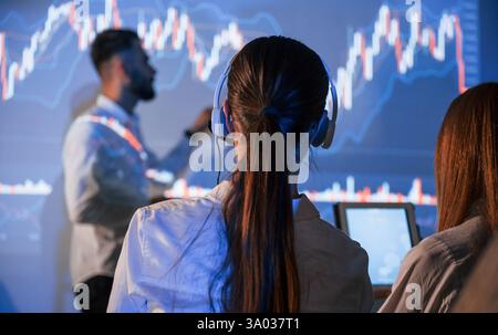 Blick von hinten auf die Frau. Männlicher Anführer, der mit Mitarbeitern spricht und den Plan auf dem Projektor im Büro zeigt. Stockfoto