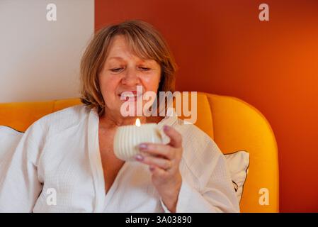 Eine ältere Frau im weißen Mantel liegt im Bett und hält eine Duftkerze. Eine reife Dame lächelt und entspannt sich im Schlafzimmer. Der Rentner schaut sich das c an Stockfoto
