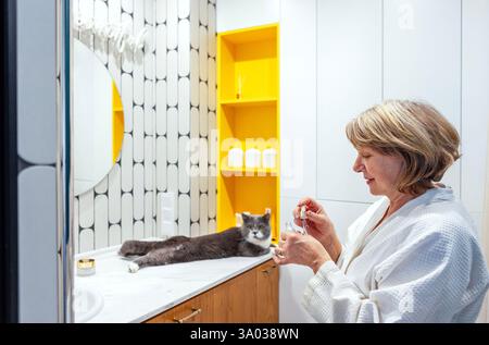Eine ältere Frau in einem weißen Hausmantel trägt Creme auf ihr Gesicht auf. Eine reife Dame kümmert sich um ihre Haut und schaut sich im Badezimmerspiegel an. Stall Stockfoto