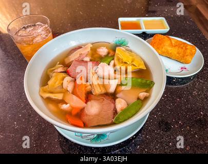 Eine köstliche Schüssel Wonton Suppe gefüllt mit Knödeln, BBQ Schweinefleisch, Huhn, Garnelen und frischem Gemüse. Serviert mit einem Eierbrötchen, eingetauchten Soßen und einem Re Stockfoto