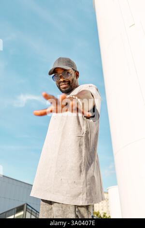 Stilvoller junger afroamerikanischer Mann mit lässiger Streetwear, Sonnenbrille und Hut, während er draußen steht und die Hand gegen den hellblauen Himmel streckt Stockfoto