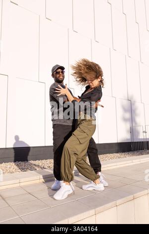Stilvolles Paar, das gemeinsam im Freien vor einer modernen weißen Wand tanzt, lässige Streetwear, Energie, Leidenschaft und urbanen Lifestyle durch dynamischen Mov trägt Stockfoto