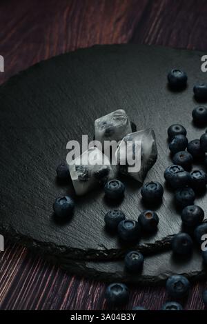 Frische Heidelbeeren und Heidelbeeren in Eiswürfeln auf einem schwarzen Steintablett auf dem Holztisch verstreut Stockfoto