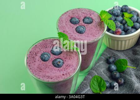 Zwei Gläser Heidelbeer-Smoothies mit Minzblättern darauf. Eine Schüssel Heidelbeeren und graues Tuch auf der Seite. Grüner Hintergrund Stockfoto