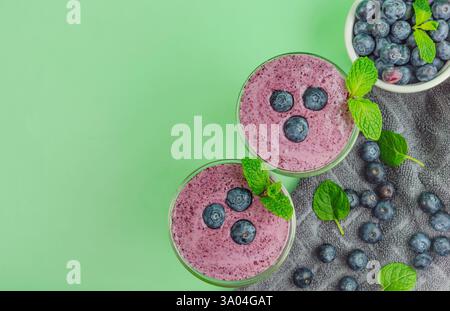 Zwei Gläser Heidelbeer-Smoothies mit Minzblättern darauf. Eine Schüssel Heidelbeeren und graues Küchentuch an der Seite. Grüner Hintergrund Stockfoto