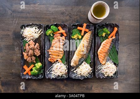 Japanische Gerichte mit verschiedenen Fleischsorten, gegrilltem Lachs, Saba, Huhn, Schweinefleisch und Gemüse auf schwarzem Keramiktablett auf Holztisch Stockfoto