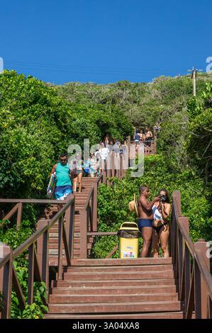 Touristen, die eine Zickzack-Holztreppe, umgeben von üppigem Grün, hinunter zum Strand Pontal do Atalaia unter dem hellen Sommermorgen sonnigen Tag hinabsteigen. Stockfoto