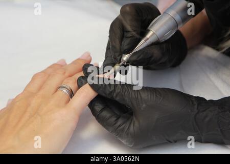 Maniküre mit schwarzen Handschuhen mit elektrischer Nagelfeile, um die Nagelhaut am Finger der Frau im Schönheitssalon zu entfernen Stockfoto