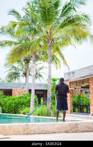 Mann, der den Swimmingpool mit Palmen im Hintergrund reinigt. Poolreinigung im tropischen Hotelresort. Luxusvilla im Freien. Hotel in Sansibar Stockfoto