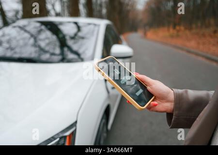 Mit Smartphone in der Hand. Ladevorgang. Die Frau steht draußen in der Nähe ihres Elektroautos auf der Straße. Stockfoto