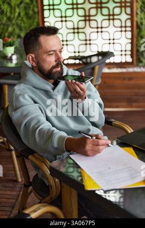 Ernsthafter junger Erwachsener in legerer Kleidung im Café nimmt Audiobotschaften auf Stockfoto