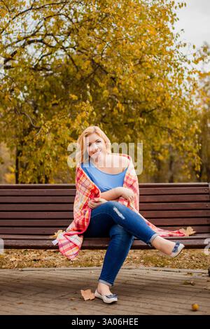 Blonde Frau in Übergröße in Jeans und blauem Hemd, die im Herbstpark mit einer Decke auf den Schultern auf einer Bank sitzt. Stockfoto