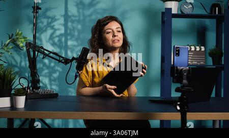 Ein technischer Experte präsentiert ein Tablet im Einstiegssegment mit niedrigen Budgets, das aus einem schwach beleuchteten Heimstudio filmt. Virale Tech-Content-Ersteller, der digitale Geräte vor Online-Publikum überprüft, Kamera B Stockfoto