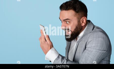 Wütender junger Manager, der das Smartphone wütend auf blauem Hintergrund anschreit Stockfoto