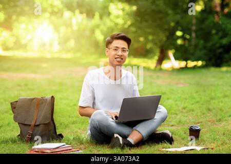 Lächelnder asiatischer Student Kerl sitzt mit Laptop im Freien, Vorbereitung für Prüfungen Stockfoto