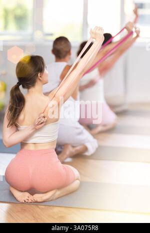 Rückansicht einer jungen, energiereichen Athletikerin, die während des Gruppenkurses im Fitnessraum mit Hilfe von Widerstandsbändern auf Matte kniend Pilates macht Stockfoto
