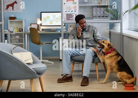 Ein langer Schuss von einem freundlichen Hund, der dem Schwarzen Mann eine Pfote gibt, der im Sessel sitzt und telefoniert, während er auf einen Termin im modernen Arztbüro wartet Stockfoto