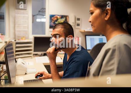 Fokussierter männlicher Gesundheitsexperte, der sich auf den Ellenbogen stützt, während er Desktop-PC mit weiblicher Kollegin in der Klinik verwendet Stockfoto