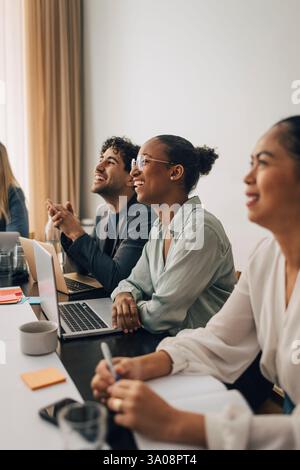 Glücklicher junger Unternehmer, der eine Brille trägt und mit männlichen und weiblichen Mitarbeitern im Besprechungsraum im Büro neben dem Laptop sitzt Stockfoto