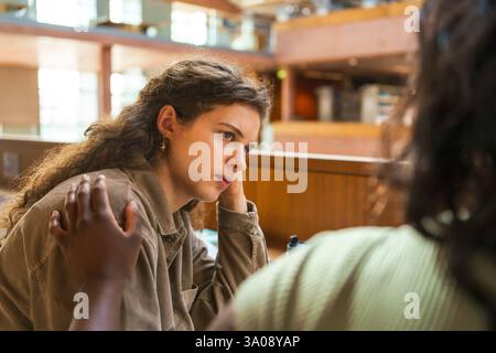 Studentin mit Hand auf der Schulter des Freundes, während sie in der Bibliothek sitzt Stockfoto