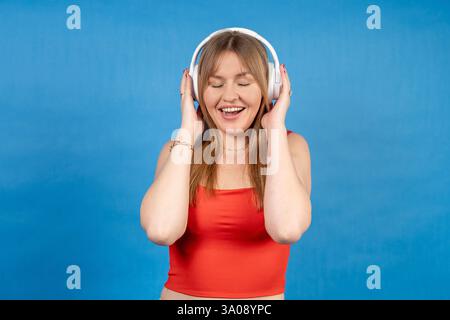 Glückliche junge Frau, die Musik mit kabellosen Kopfhörern hört, den Klang genießt und mit geschlossenen Augen auf blauem Hintergrund lächelt Stockfoto