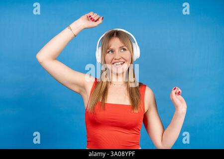 Sorglose junge Frau, die kabellose Kopfhörer trägt, genießt Musik, tanzt und lächelt auf einem leuchtend blauen Hintergrund Stockfoto