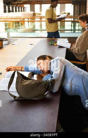 Müde Frau schläft auf dem Schreibtisch neben Studenten, die in der Bibliothek studieren Stockfoto