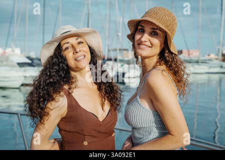 Zwei fröhliche Frauen tragen Sommerhüte und lächeln in einem Jachthafen, während sie einen sonnigen Urlaubstag am Meer genießen. Perfektes Konzept für Sommerreisen und Freundlichkeit Stockfoto