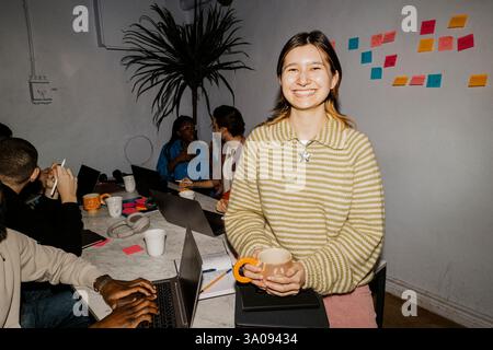 Porträt einer lächelnden Unternehmerin, die Kaffeetasse hält, während sie im Büro auf dem Tisch sitzt Stockfoto