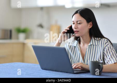 Asiatische Frau, die ihren Laptop überprüft, telefoniert in einer Küche zu Hause Stockfoto