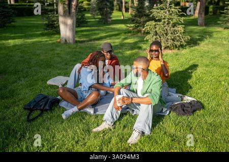 Multikulturelle Freunde, die sich auf dem Gras des Parks entspannen, Picknick genießen, plaudern und gemeinsam Drinks trinken Stockfoto