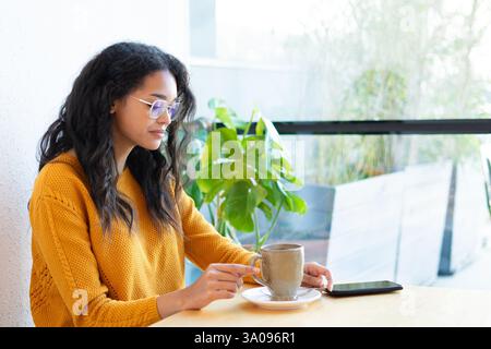 Junge lateinfrau genießt eine entspannende Kaffeepause, während sie ihr Smartphone in einem hellen und gemütlichen Café durchsucht Stockfoto