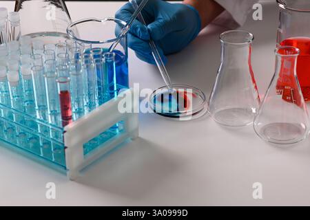 Mit Handschuhen versehene Hand mit einer Pipette: Geben Sie einen Tropfen Reagenz auf die Petrischale mit buntem Wirbel, Bechergläser und Reagenzgläser im Hintergrund auf weißem Labortisch. Stockfoto