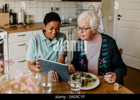 Hochwinkelansicht einer lächelnden Hausmeisterin, die der älteren Frau einen Tablet-PC zeigt, der am Tisch in der Küche frühstückt Stockfoto