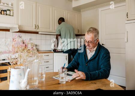Senior Mann löst Kreuzworträtsel in der Zeitung, während männliche Krankenschwester zu Hause in der Küche arbeitet Stockfoto