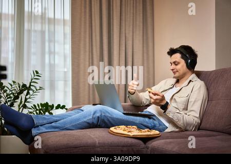 Junger Mann, der auf dem Sofa sitzt und Pizza isst, während er einen Videoanruf mit einem Freund auf dem Laptop hat Stockfoto
