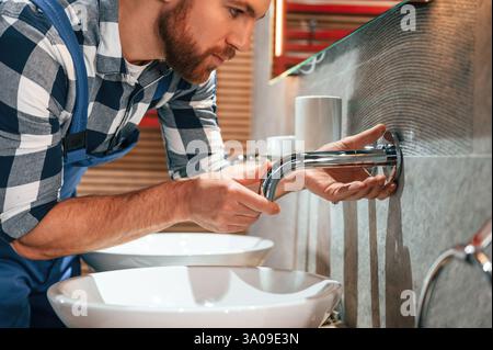 Nahansicht. Wasserhahn wird befestigt. Klempner in blauer Uniform arbeitet im Badezimmer. Stockfoto
