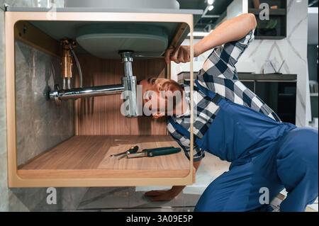 Beschäftigt mit der Reparatur des Siphons. Klempner in blauer Uniform arbeitet im Badezimmer. Stockfoto